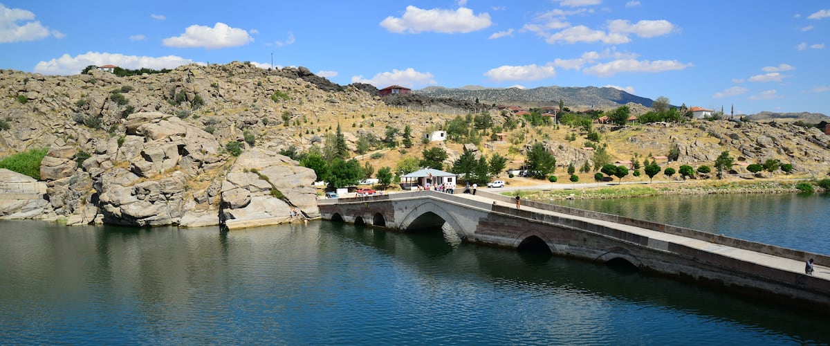 A stone bridge over a calm river, surrounded by rocky terrain and distant mountains.