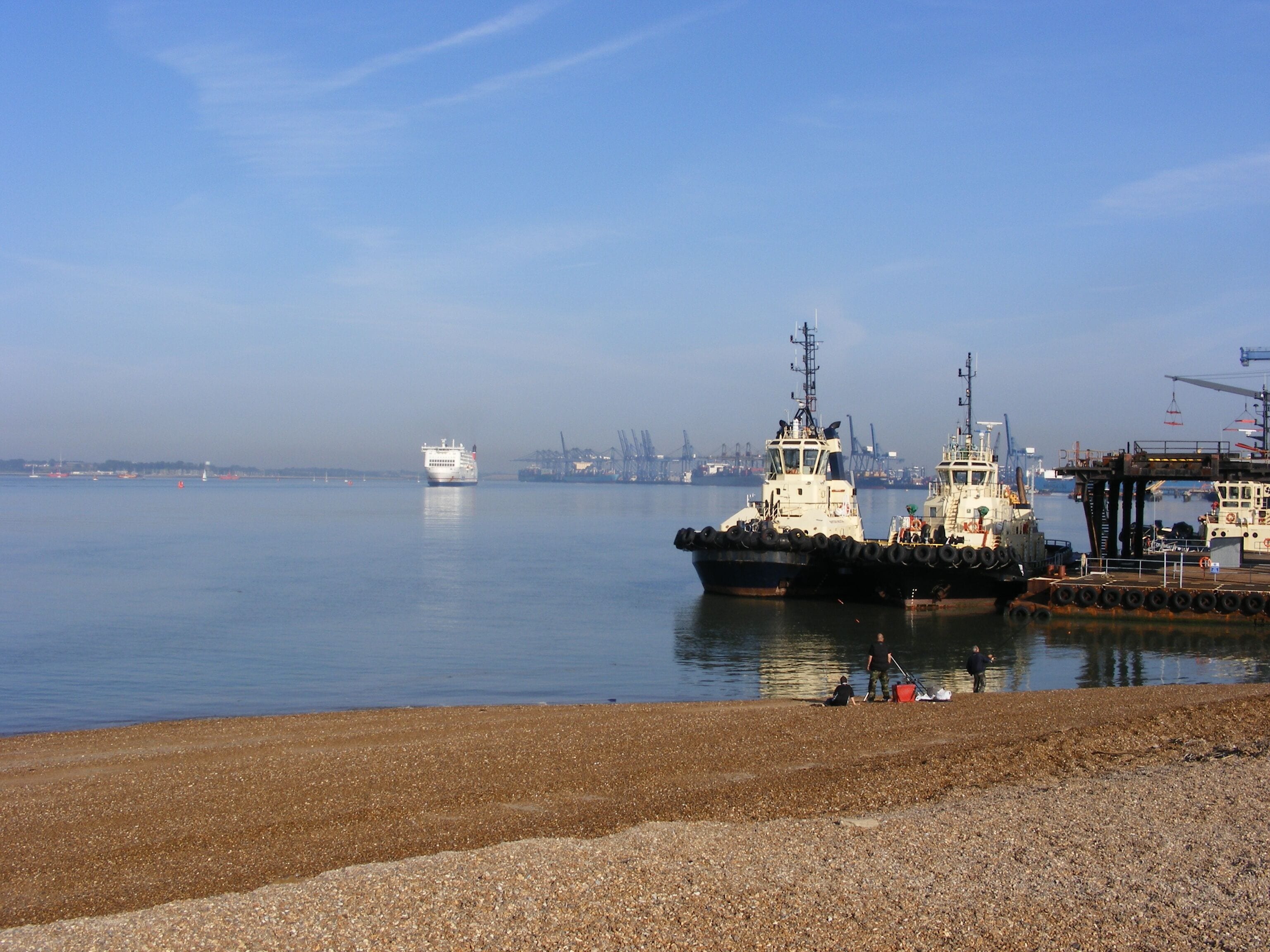 A harbor with two large ships docked, a calm sea, and industrial cranes in the background.