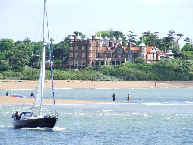 A sailboat on calm waters, a sandy beach, and a large red brick building with turrets.