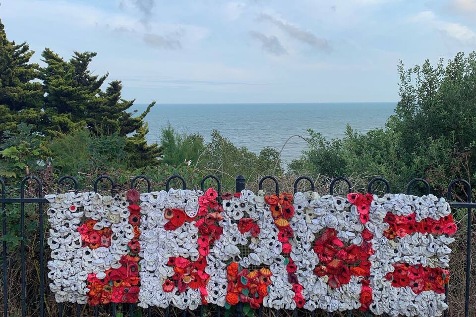 Une pancarte en crochet représentant le mot « PAIX », ornée de fleurs rouges et blanches, placée sur un fond de ciel nuageux et offrant une vue sur la mer.