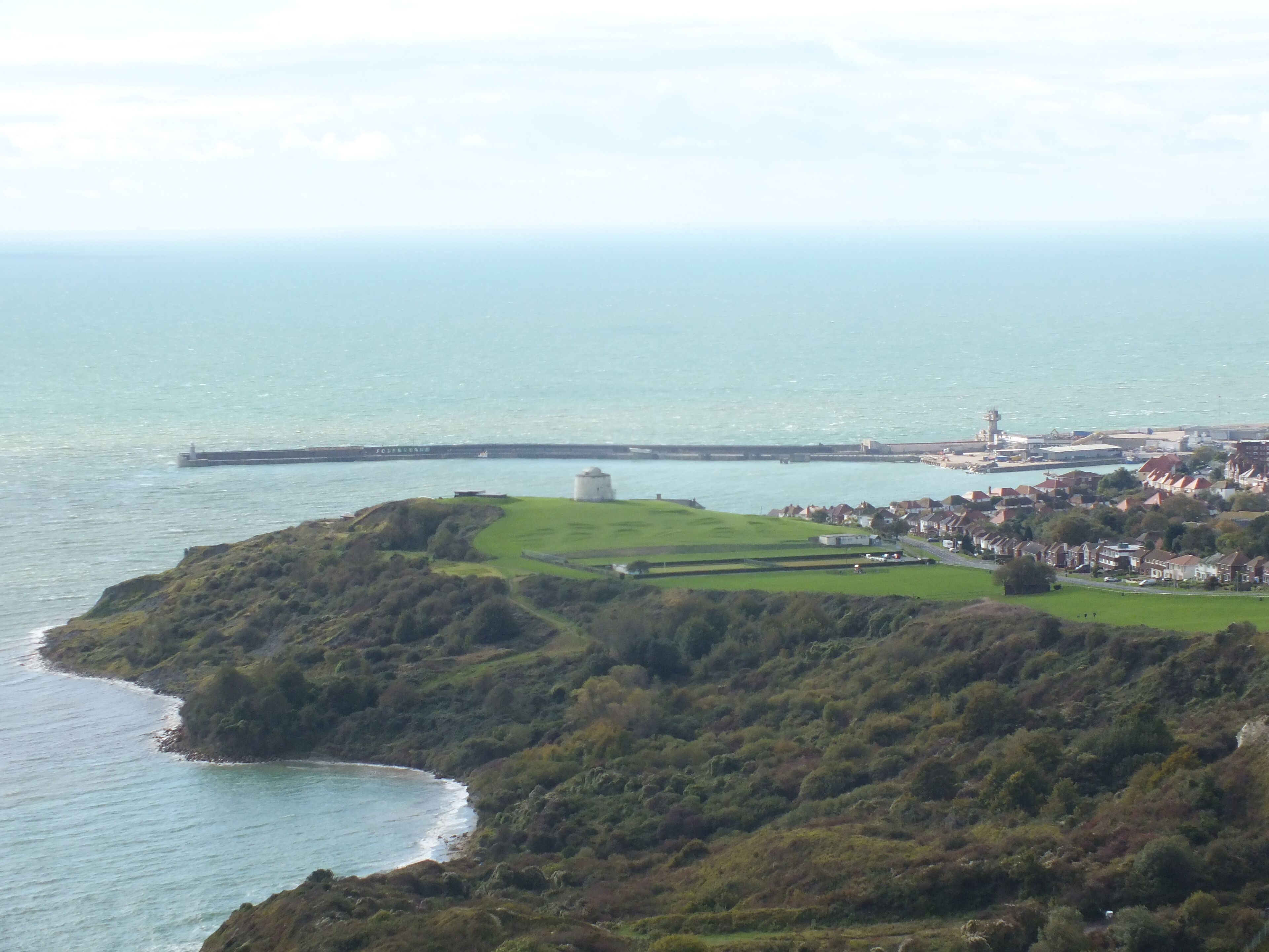 A coastal landscape with a lighthouse, a pier, and a town with houses.