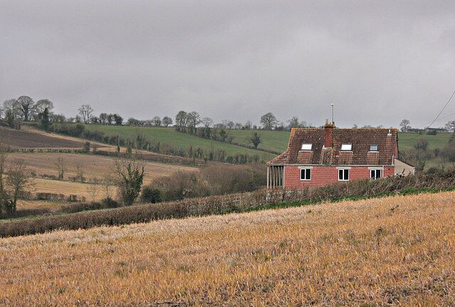 A red brick house with a tiled roof in a rural landscape with rolling hills and fields.