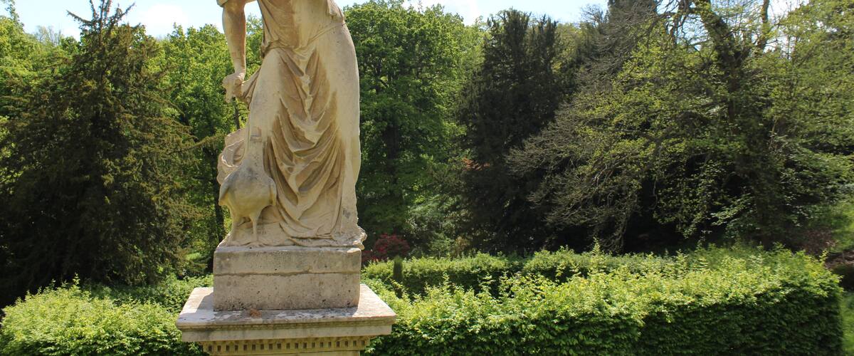 A classical statue in a lush garden with dense greenery and trees in the background.