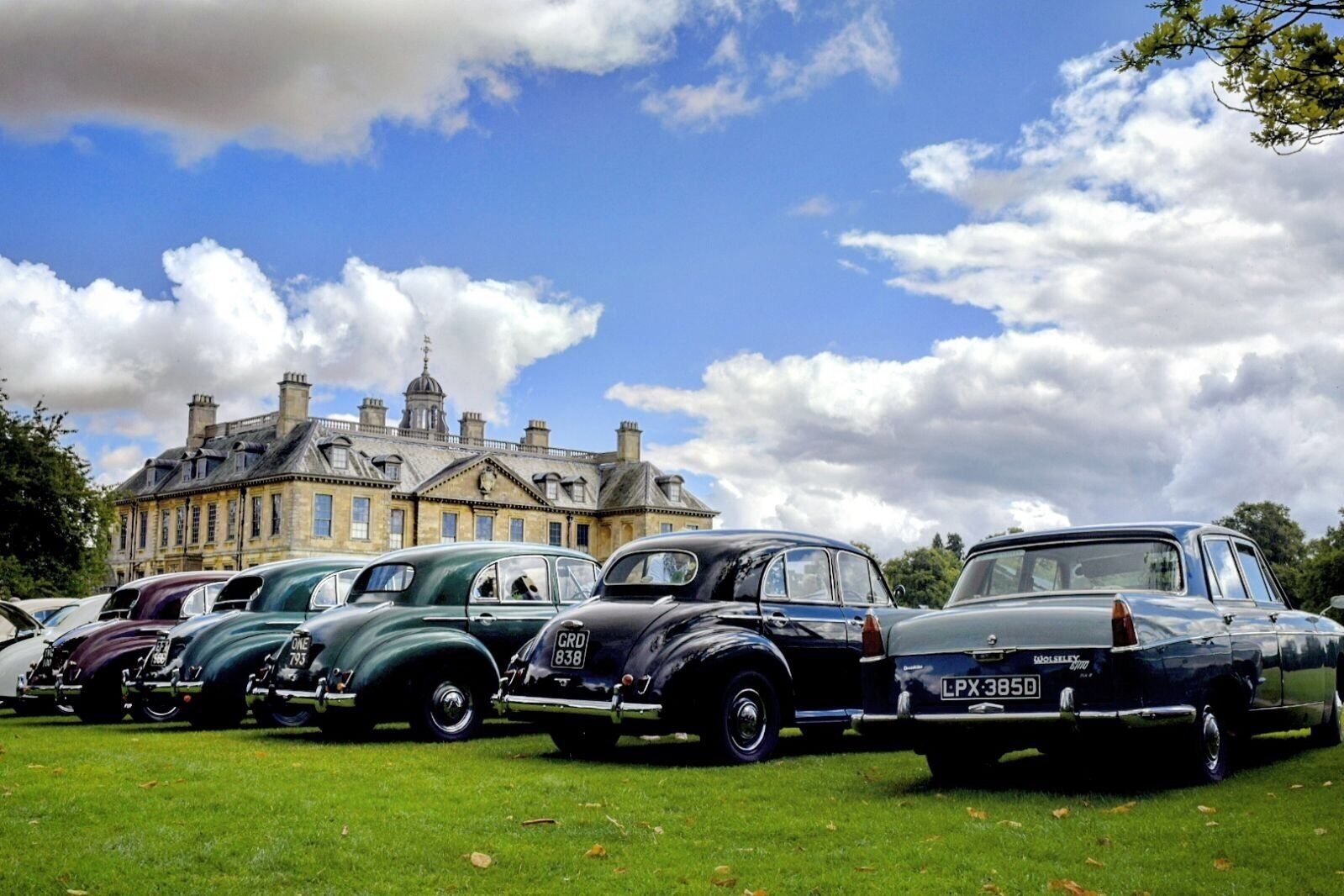 A row of vintage cars parked on a grassy field with a large historic building in the background.