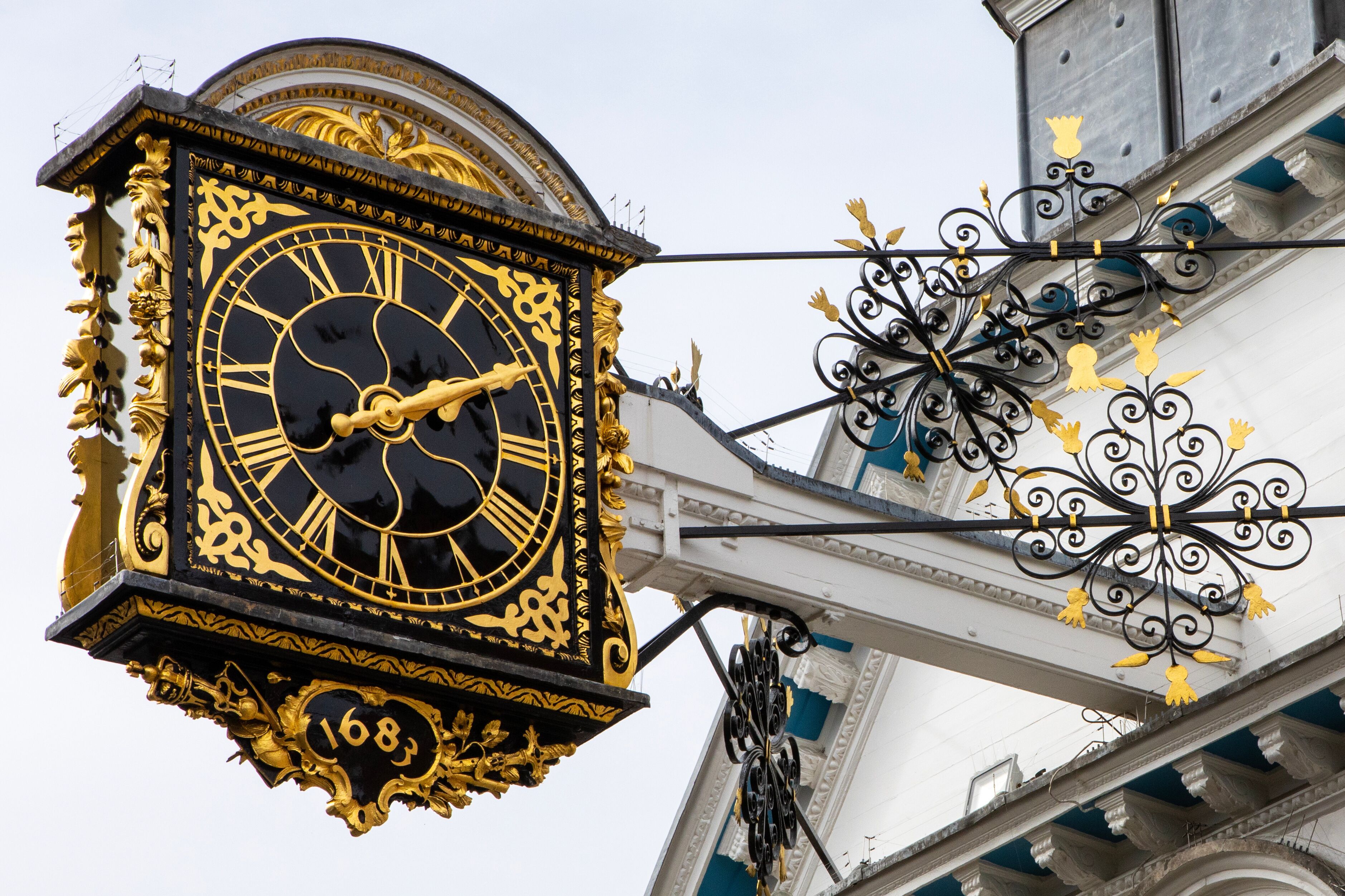 Un reloj grande y ornamentado con números romanos, adosado a un edificio con detalles metálicos decorativos.