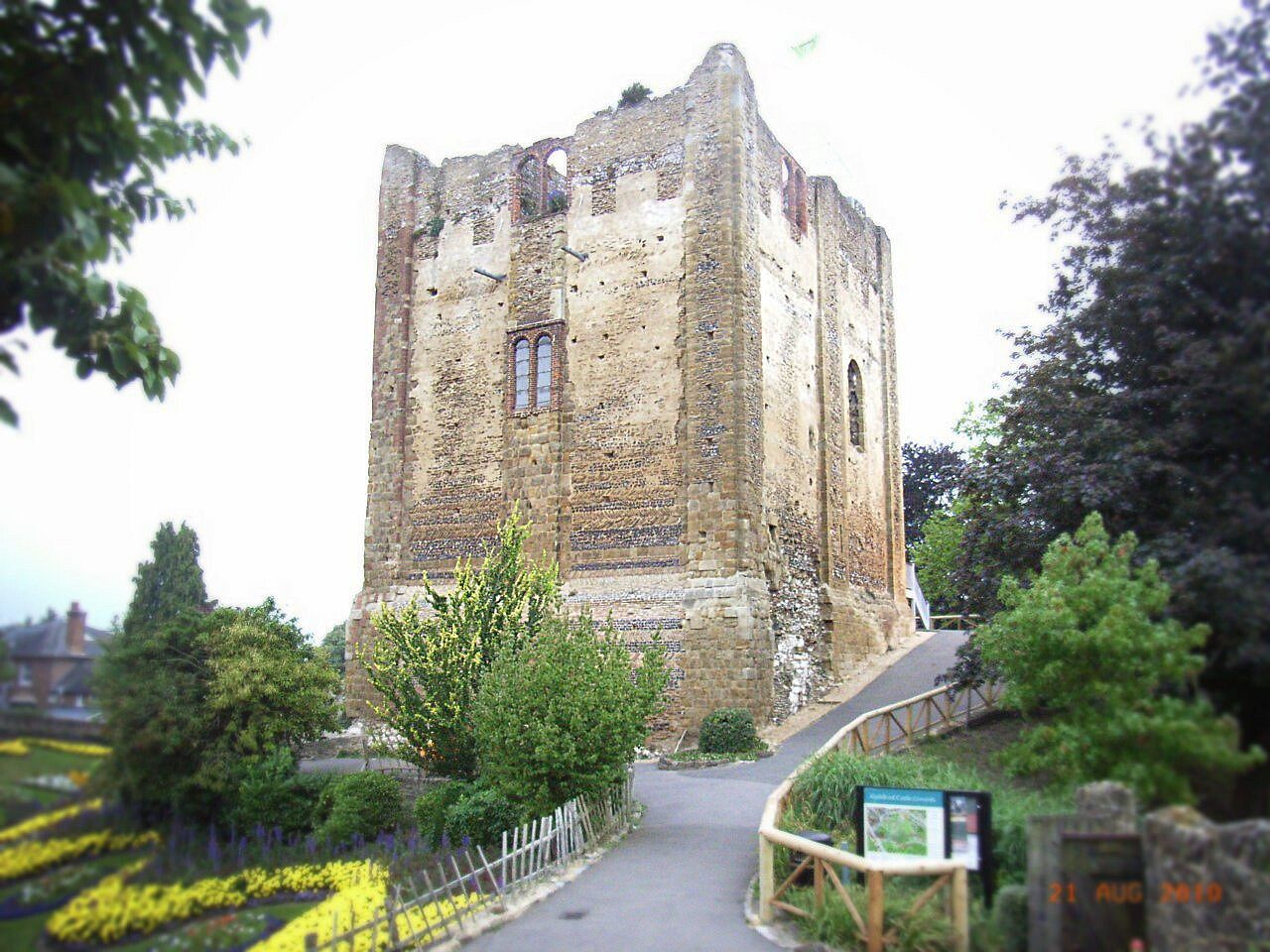 A historic stone tower with a narrow entrance pathway, surrounded by greenery and a paved road.