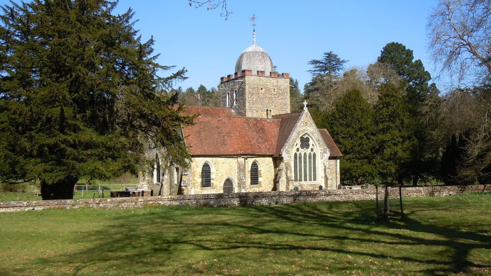 A historic stone church with a red-tiled roof, surrounded by a grassy field and tall trees.