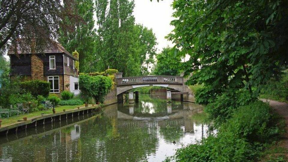 A canal with a stone bridge, lush greenery, and a house on the left.