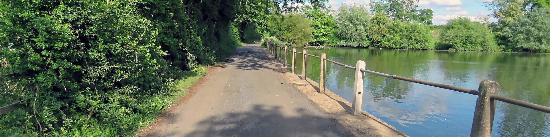 The access road running northeast away from St Mary's Parish Church, with concrete post and scaffolding barrier against an estate mid-19th-century artificial fishing lake, in the civil parish of Matching, in Essex, England. Camera: Canon PowerShot SX60 HS Software: File lens-corrected, optimized, perhaps cropped, with DxO OpticsPro 11 Elite, and likely further optimized with Adobe Photoshop CS2.