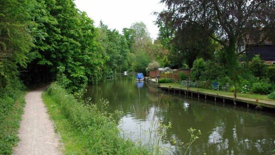 A narrow waterway with a path on the left, surrounded by dense greenery and a dock on the right.
