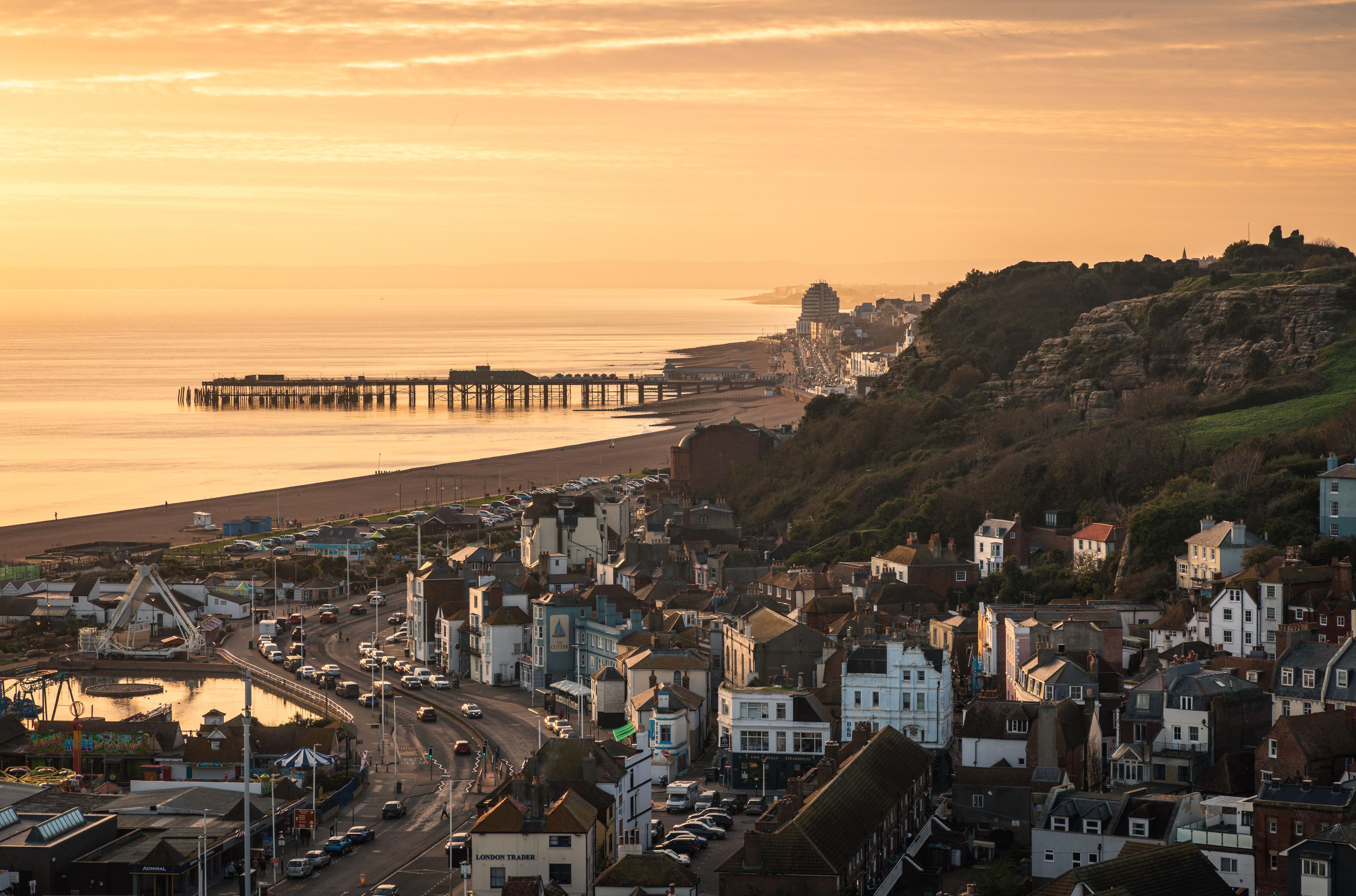 A coastal town with a pier extending into the sea, surrounded by buildings and hills.