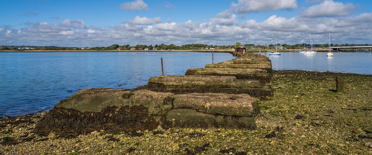 A stone pier extending into a calm body of water, surrounded by a rocky shoreline and a distant bridge.
