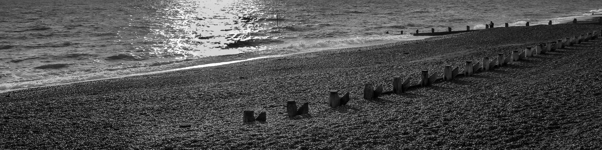 Uma praia de pedrinhas com uma fileira de estacas de madeira e um mar agitado sob um céu nublado.