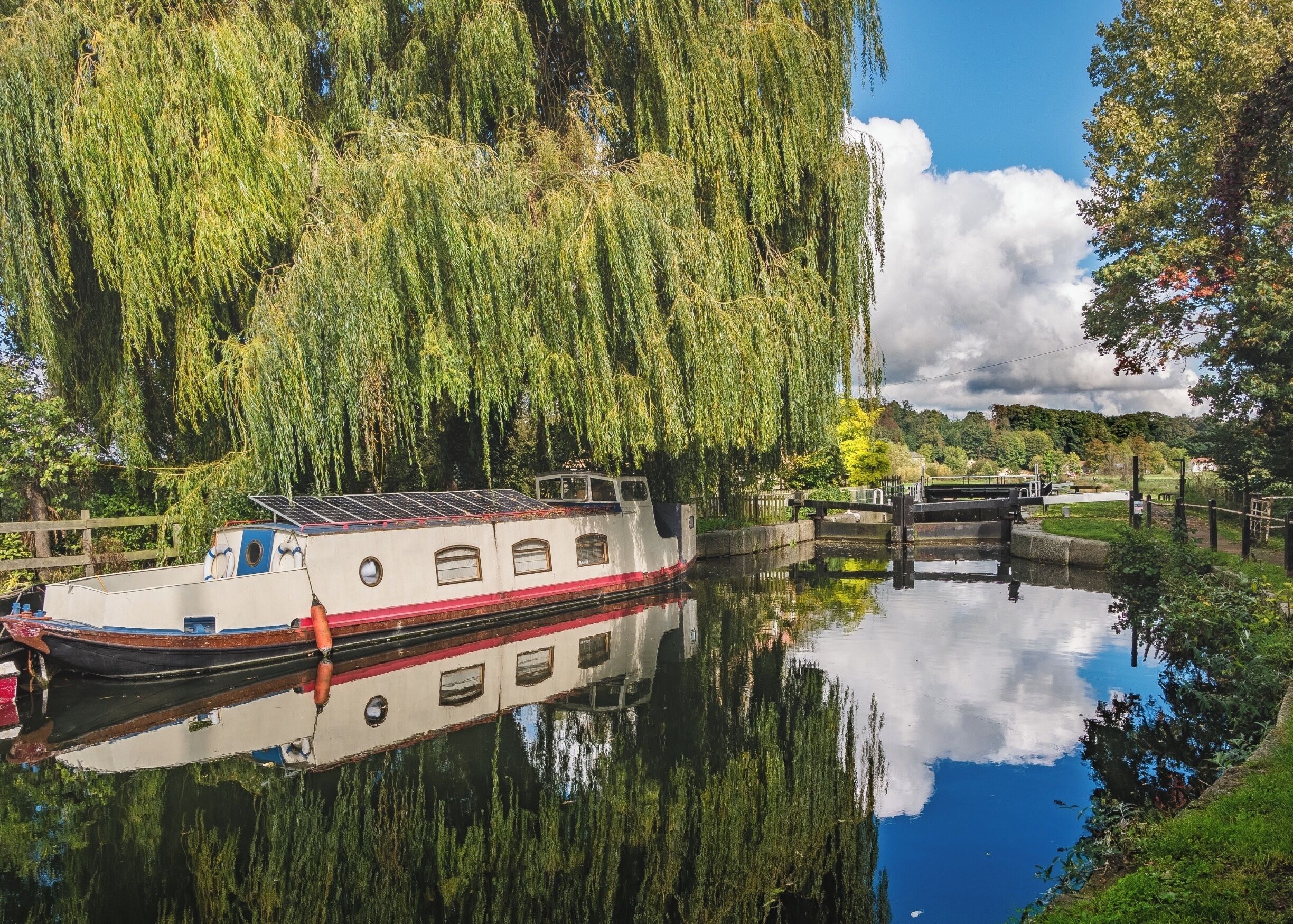 A narrowboat docked by a weeping willow tree, a lock system, and a calm river reflecting the surroundings.