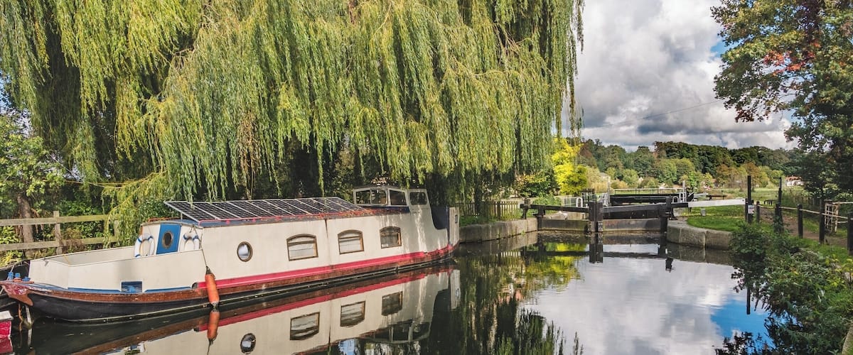 A narrowboat docked by a weeping willow tree, a lock system, and a calm river reflecting the surroundings.