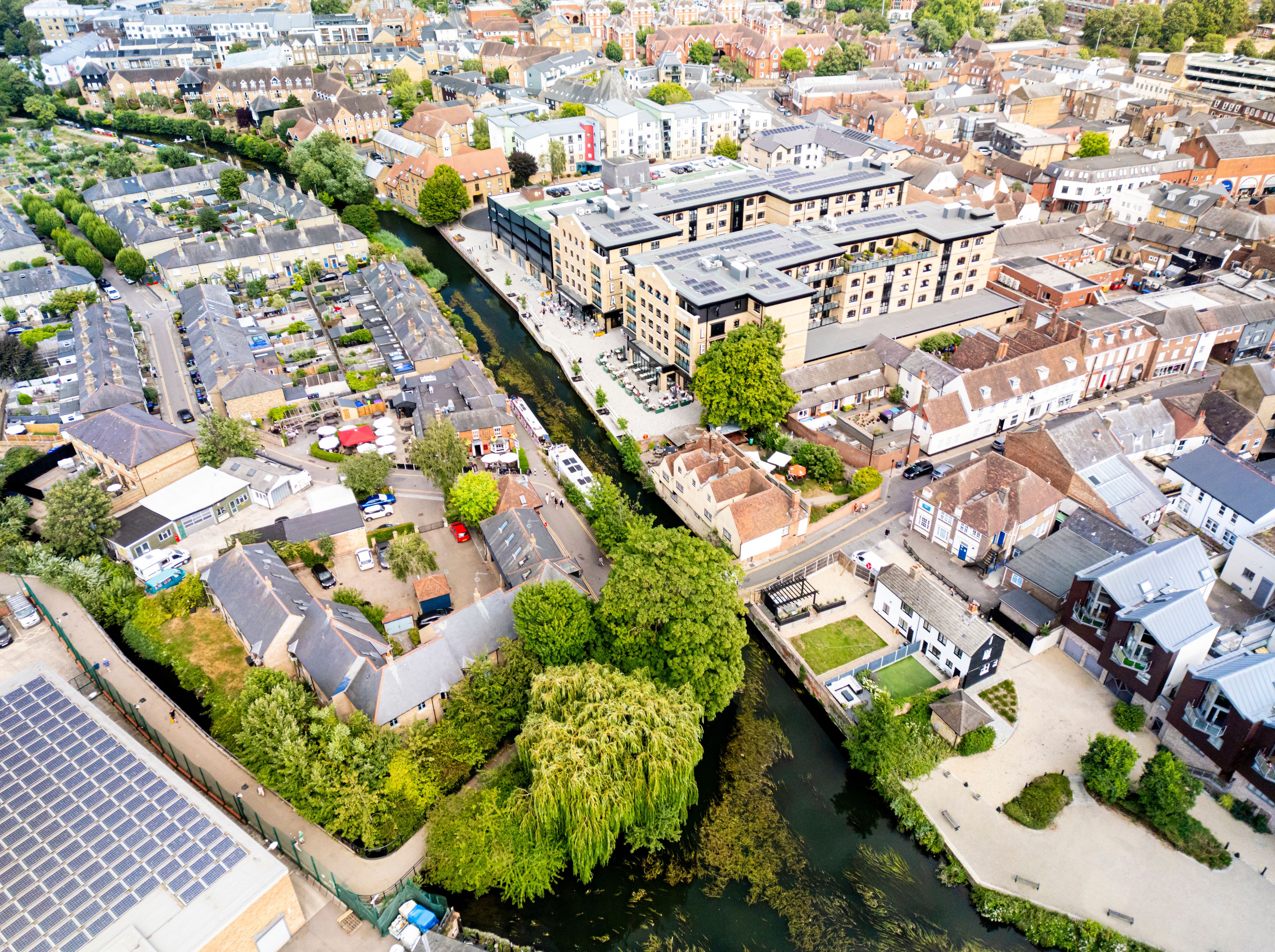 Vista aérea de un pueblo ribereño con viviendas densas, una zona verde destacada con árboles y un río claro.