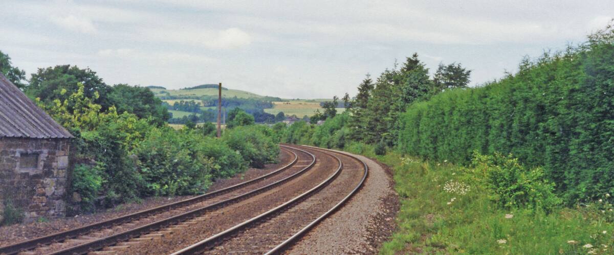 Railway tracks curving through a rural landscape with greenery and a stone building.