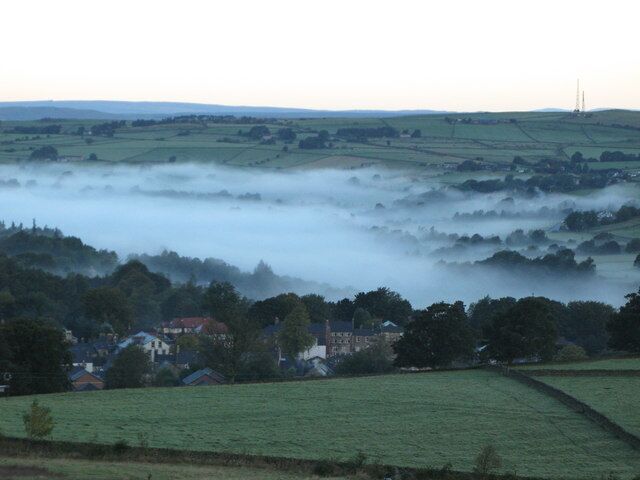 A misty rural landscape with rolling green fields, scattered houses, and a distant power line tower.