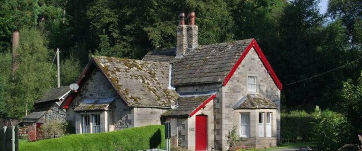 A stone house with a red door and a red roof, surrounded by greenery and a winding road.