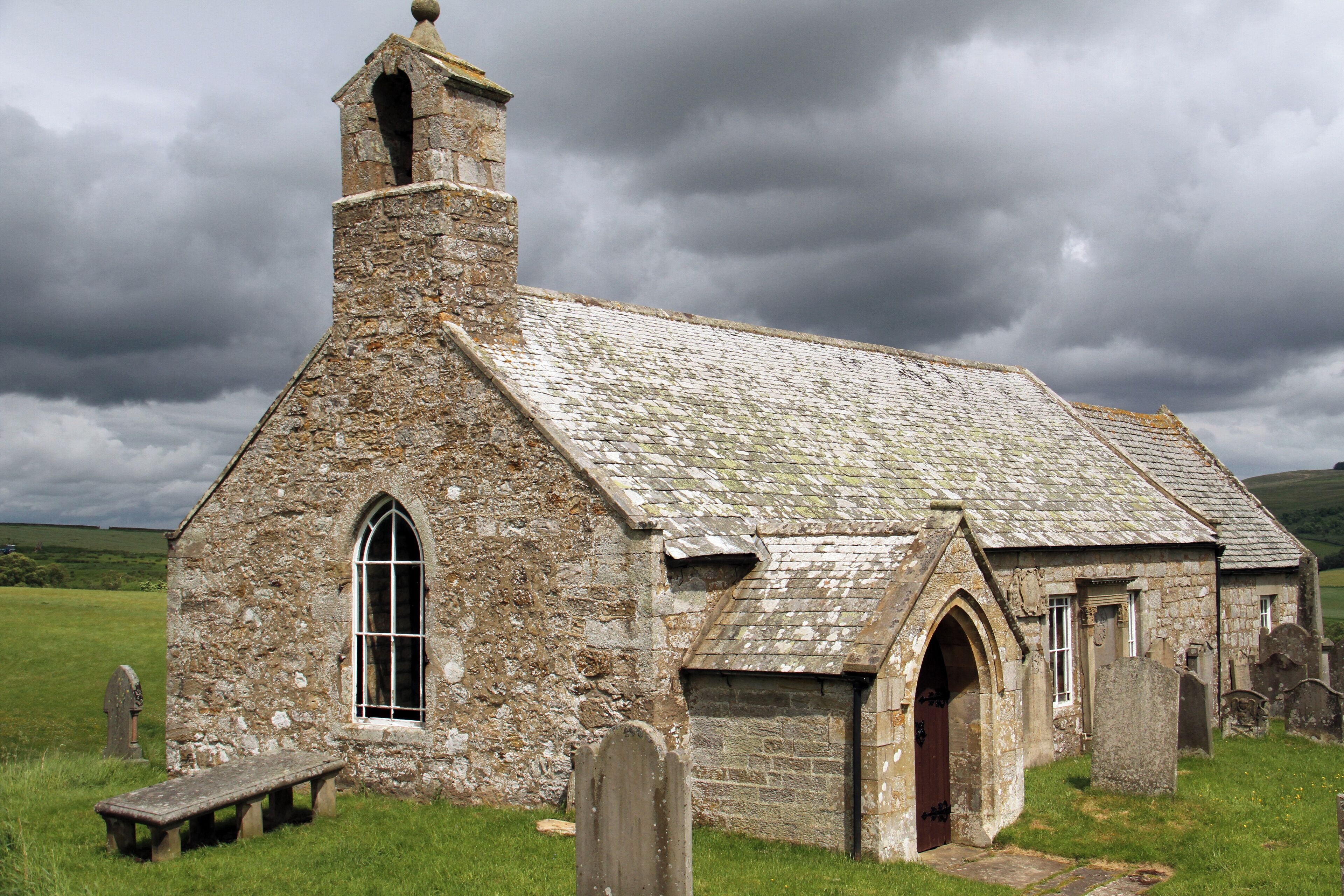 A stone church with a bell tower and arched windows in a grassy field.
