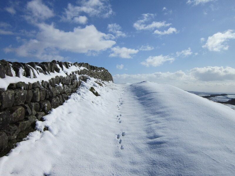 A snow-covered path with footprints leading to a stone wall under a clear sky.