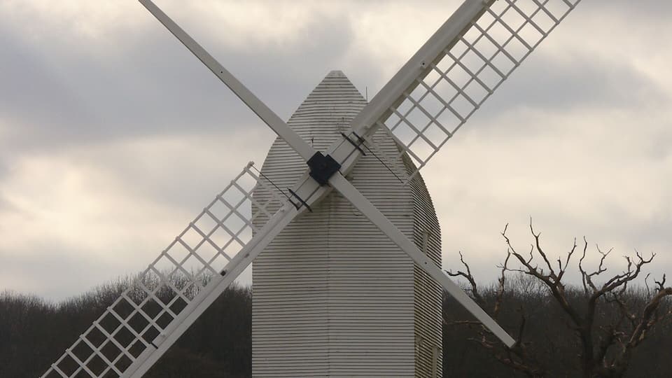 A windmill with a white lattice structure and a dark body, set against a cloudy sky and surrounded by trees and a fence.