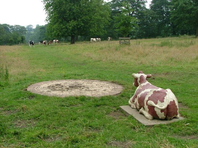 Escultura de vaca em um campo gramado com um padrão circular no centro e árvores ao fundo.