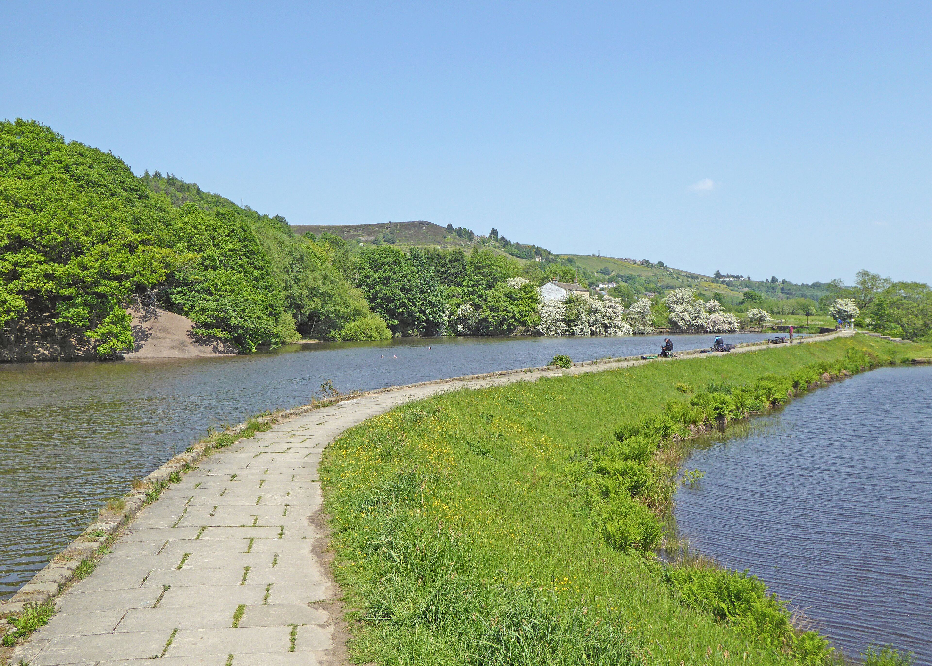 A river with a paved walkway, lush greenery, and a few people in the distance.