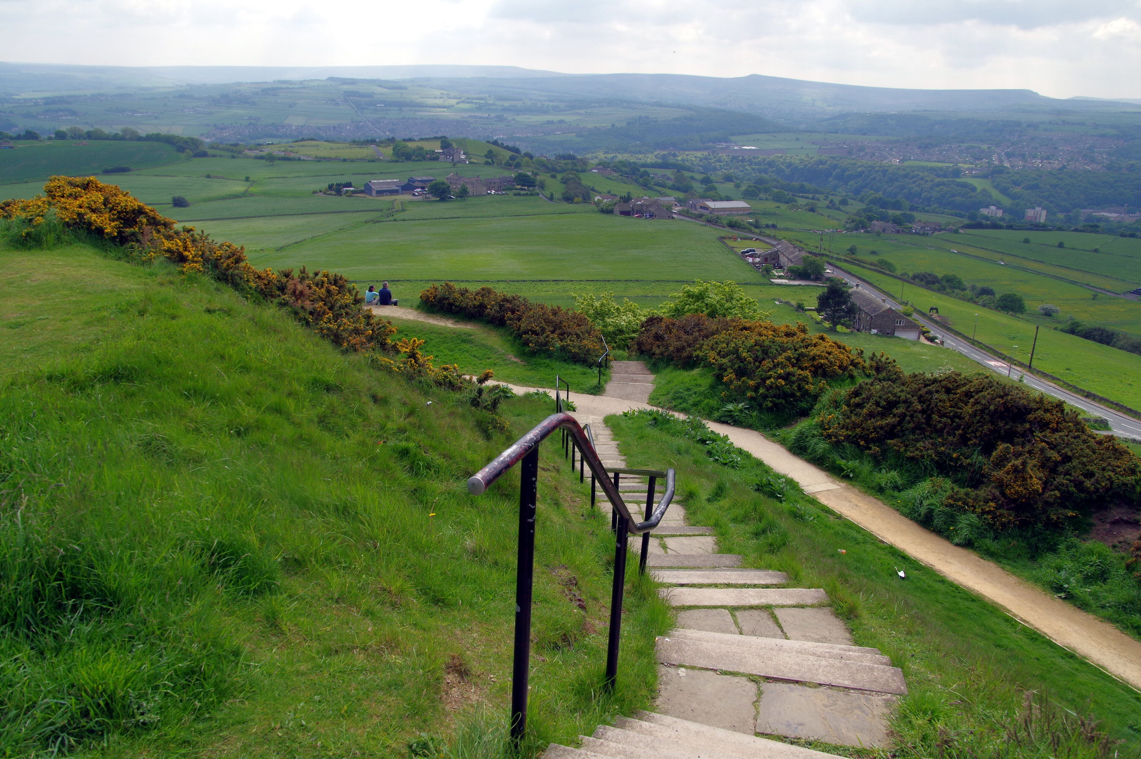 A stone staircase leading down a grassy hill with a view of a rural landscape, including fields and distant buildings.