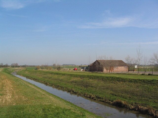 A rural landscape with a narrow water channel, a brick barn, and open fields.