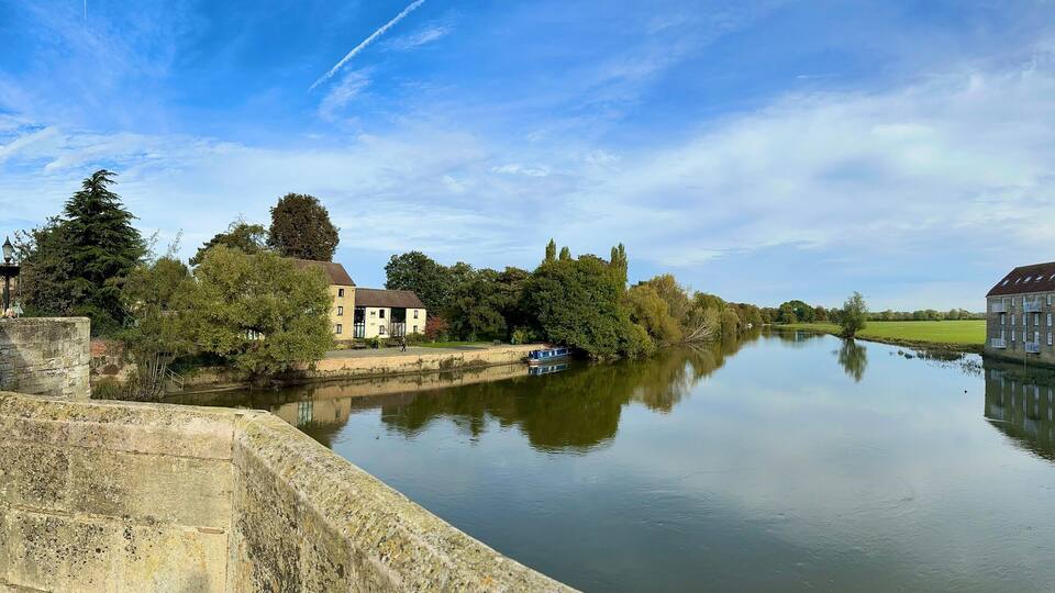 A river with a stone bridge, a large brick building with a curved roof, and a clear blue sky.