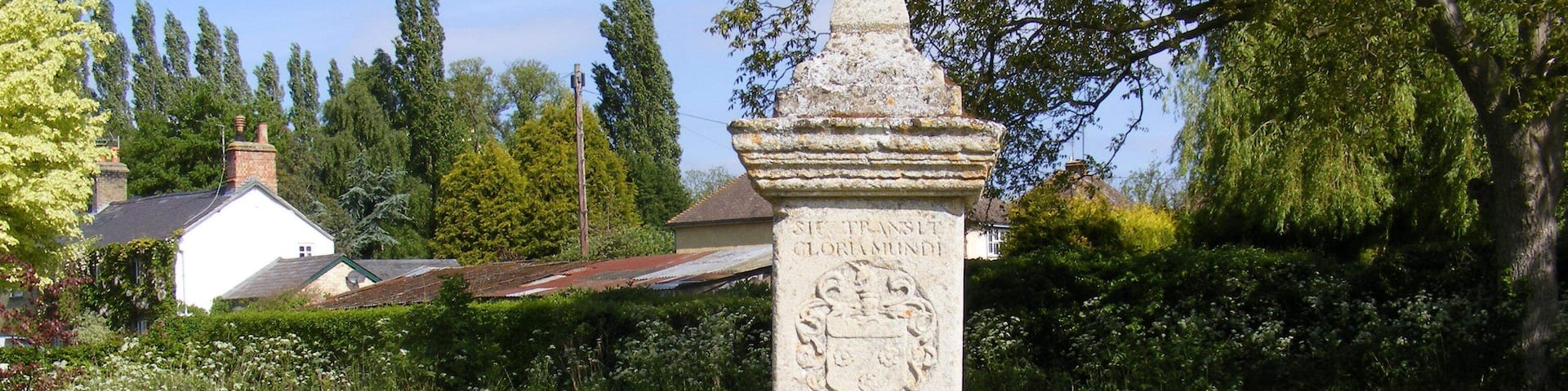 A stone monument with an inscription in a grassy area with trees and a house in the background.