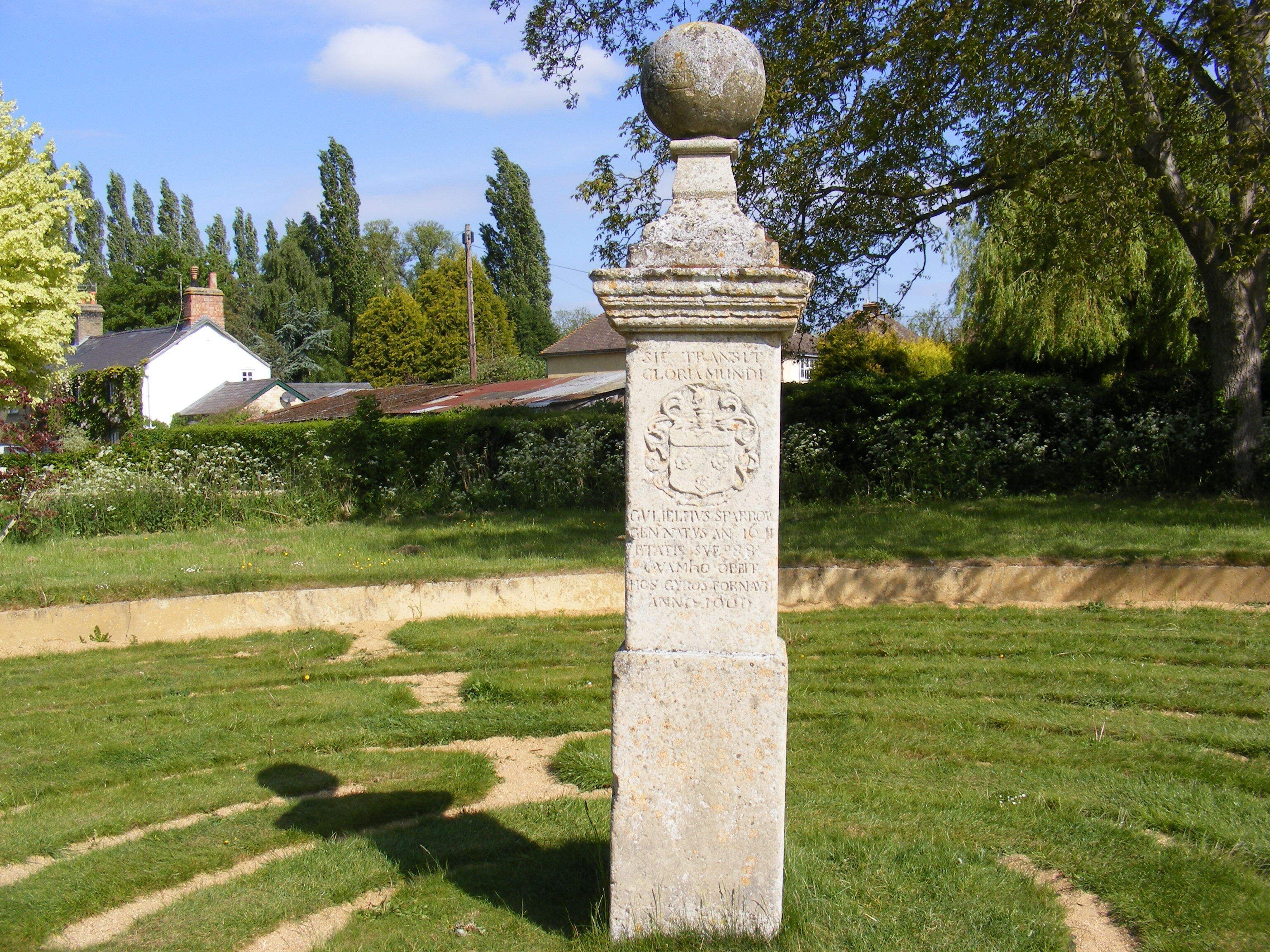 A stone monument with an inscription in a grassy area with trees and a house in the background.