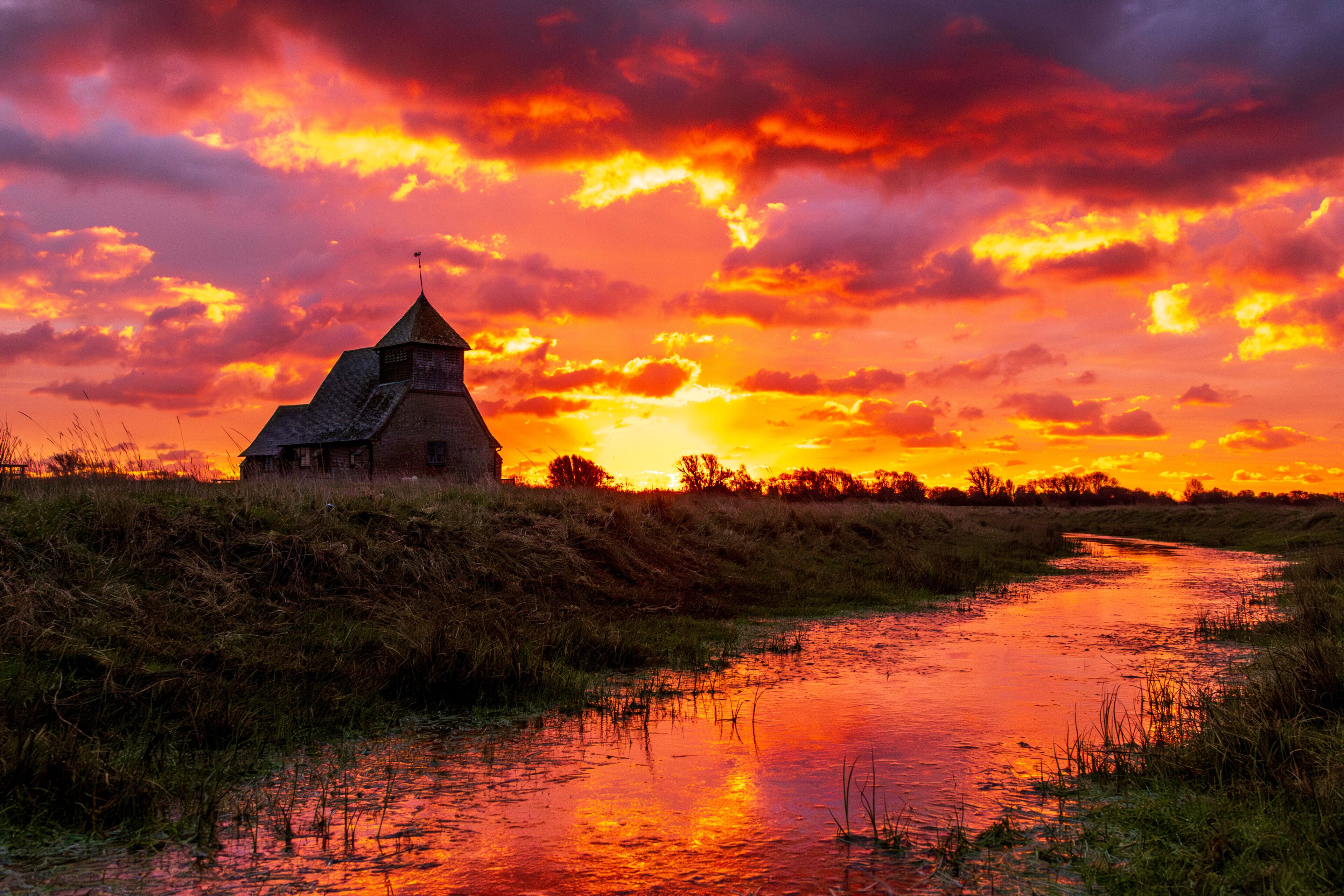 A sunset over a river with a building on a grassy bank.