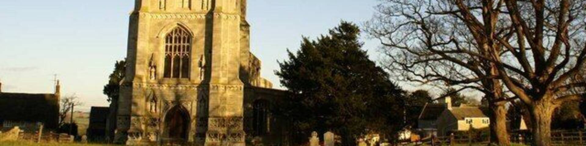 A tall church tower with a pointed spire, surrounded by a grassy field and trees.