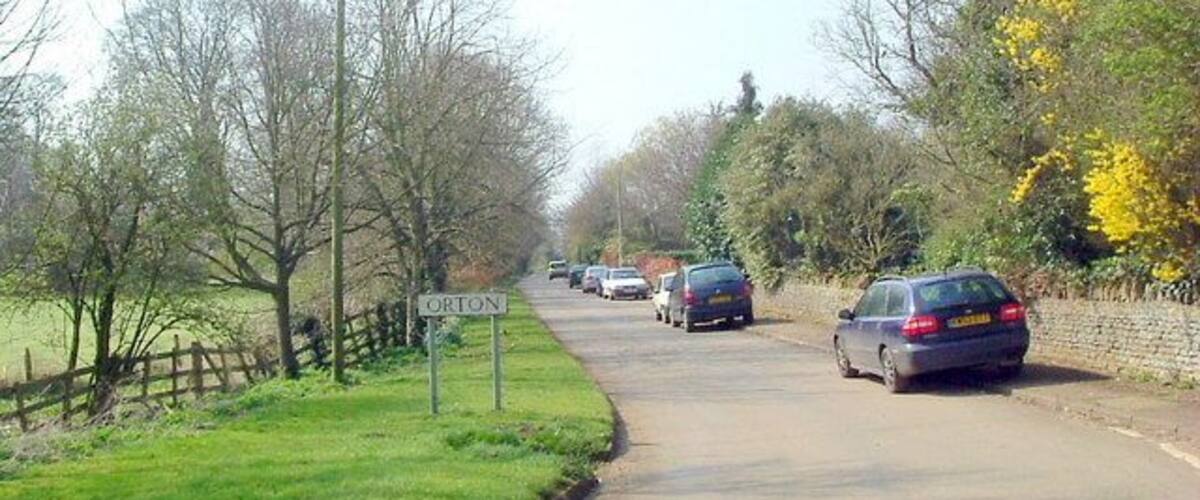 A rural road with a 'ORTON' sign, surrounded by trees and grass.