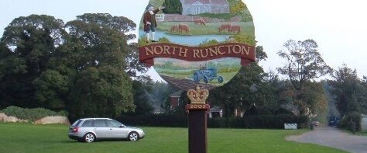 A signpost with 'North Runcott' written on it, set against a grassy field with a car in the background.