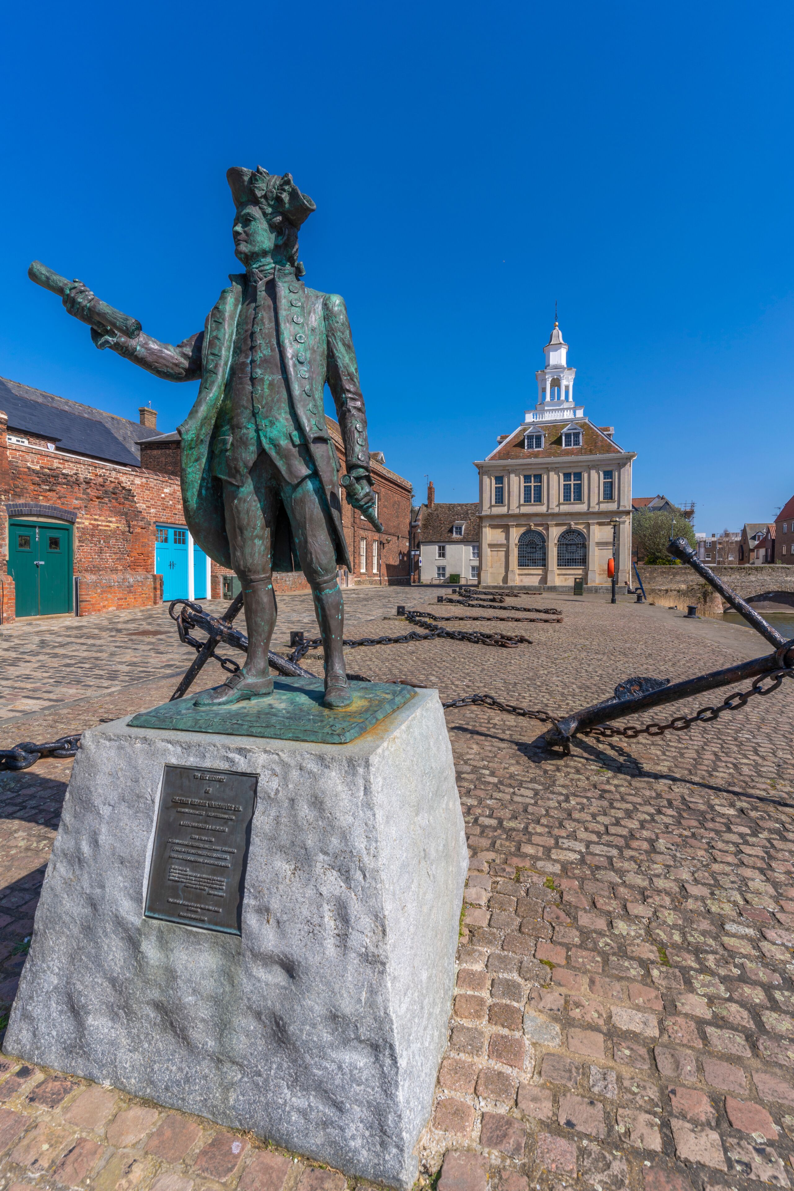 A statue of a historical figure on a pedestal, a cobblestone street, and a building with a tower in the background.