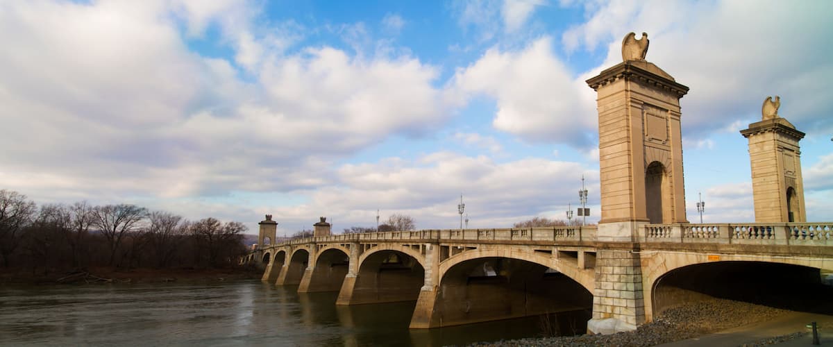 A stone bridge with multiple arches over a river, accompanied by two tall towers with statues on top.