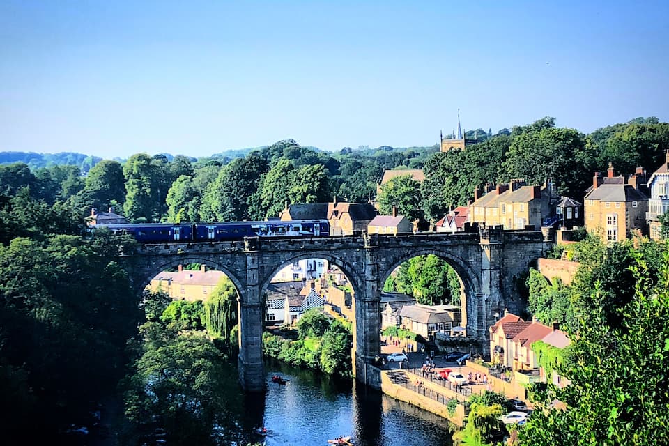 A stone bridge over a river with a train crossing, surrounded by greenery and historic buildings.