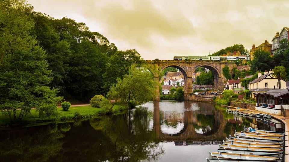 A river with boats docked on the right, a stone bridge in the background, and lush greenery on the left.