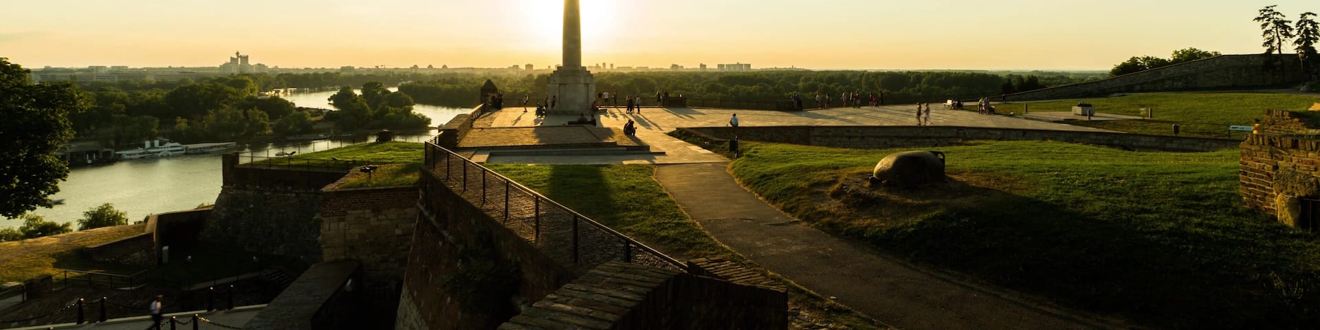 A historical monument with a tall column, a river in the background, and a park with pathways.