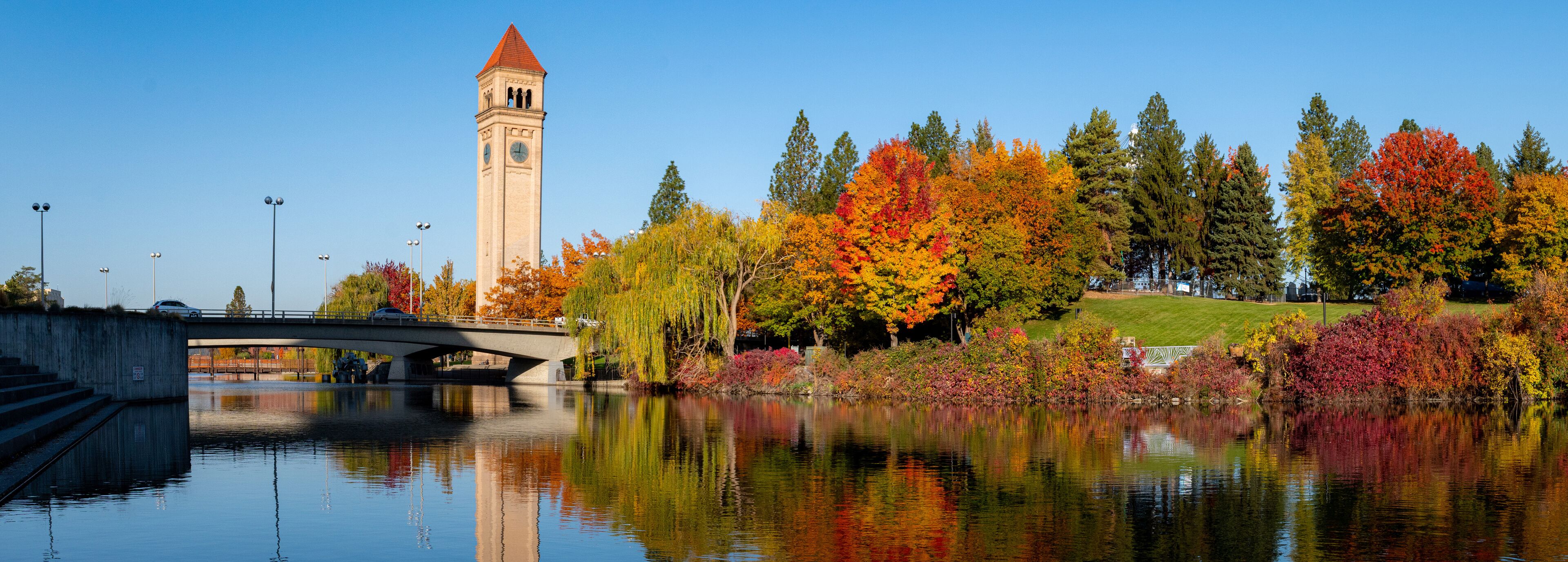 A bridge over a calm river, autumn trees with vibrant foliage, and a clock tower.