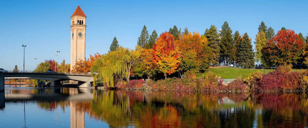 A bridge over a calm river, autumn trees with vibrant foliage, and a clock tower.