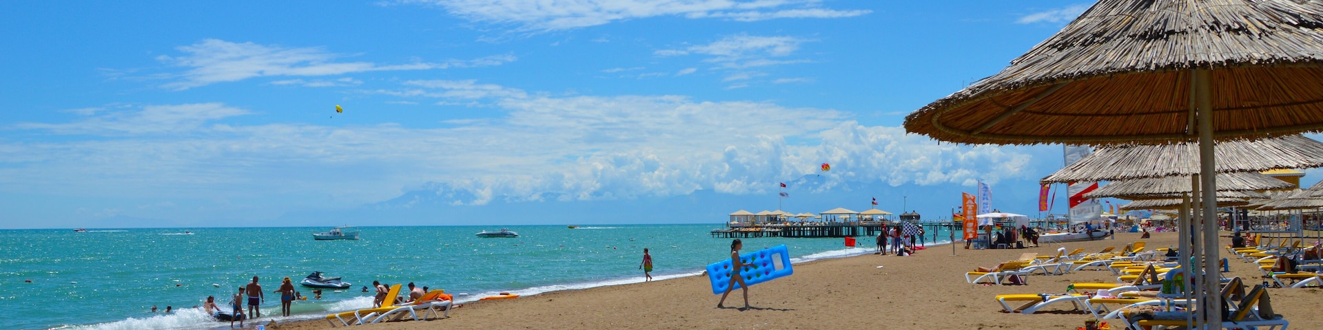 A beach with thatched umbrellas, sun loungers, and a pier in the background.
