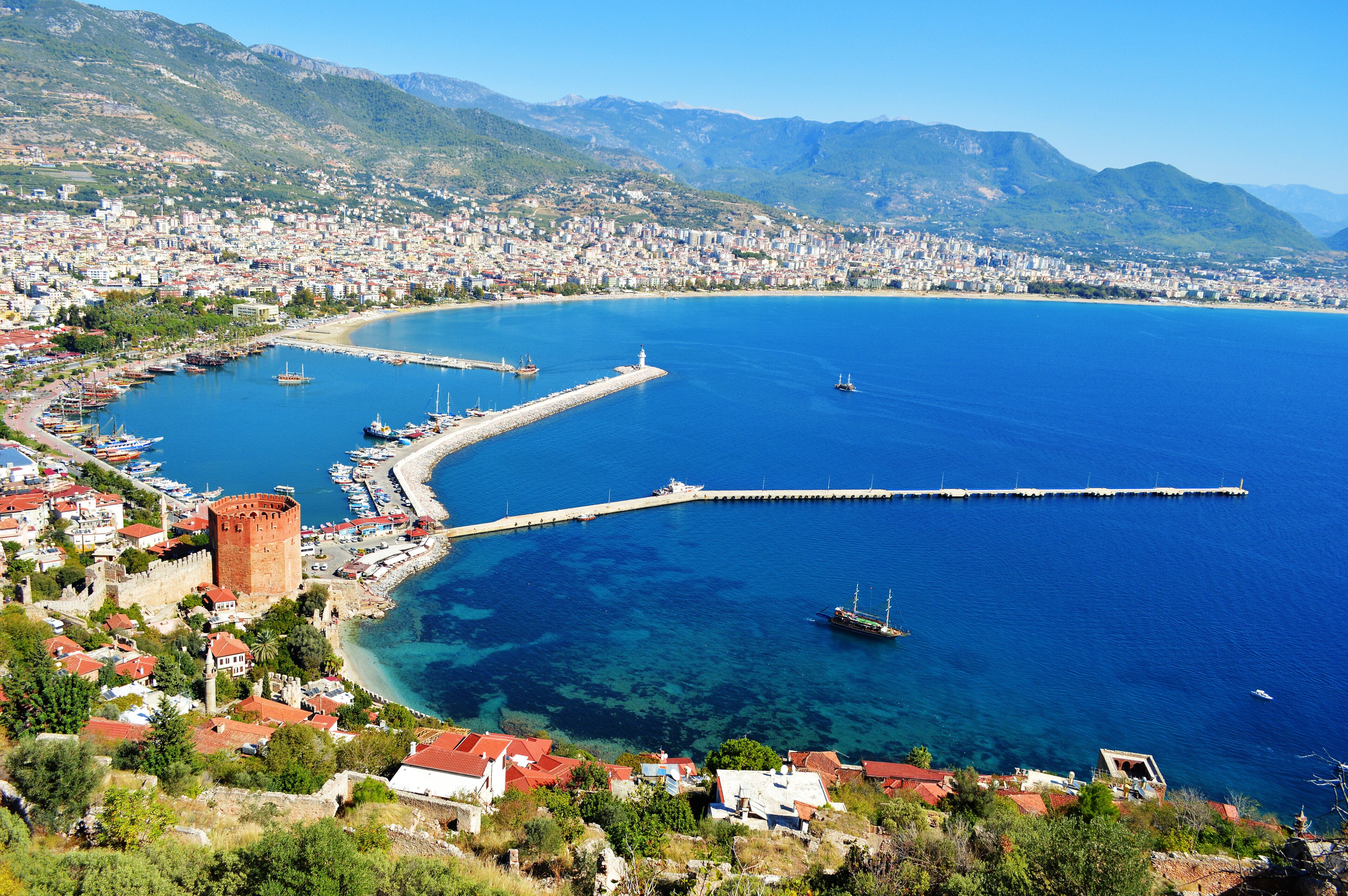 Una ciudad costera con mar azul cristalino, un muelle destacado y una torre histórica.