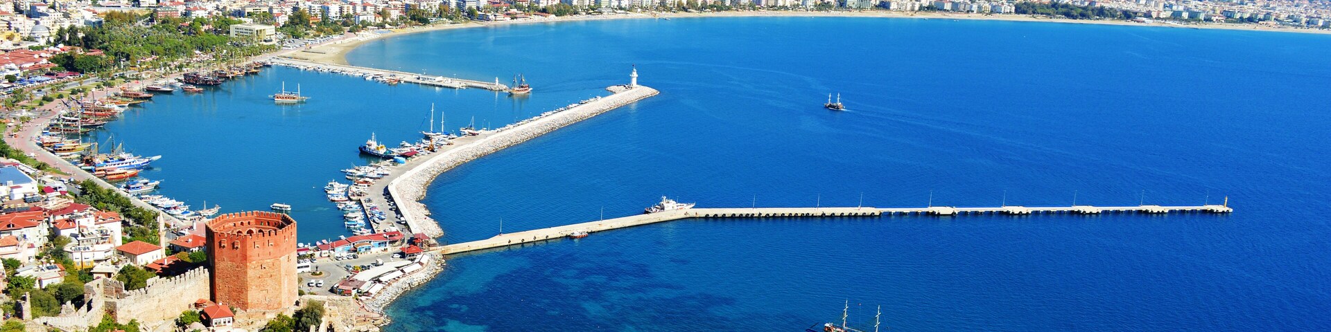 Una ciudad costera con mar azul cristalino, un muelle destacado y una torre histórica.