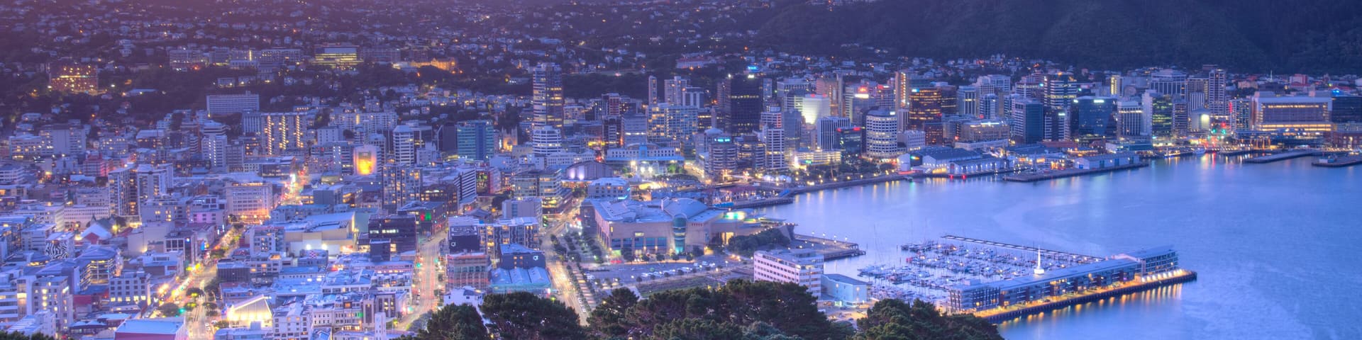 A coastal cityscape at dusk with illuminated buildings, a marina, and a backdrop of hills.