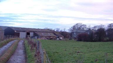 A rural landscape with a gravel path, a wooden fence, a grassy field, and farm buildings.