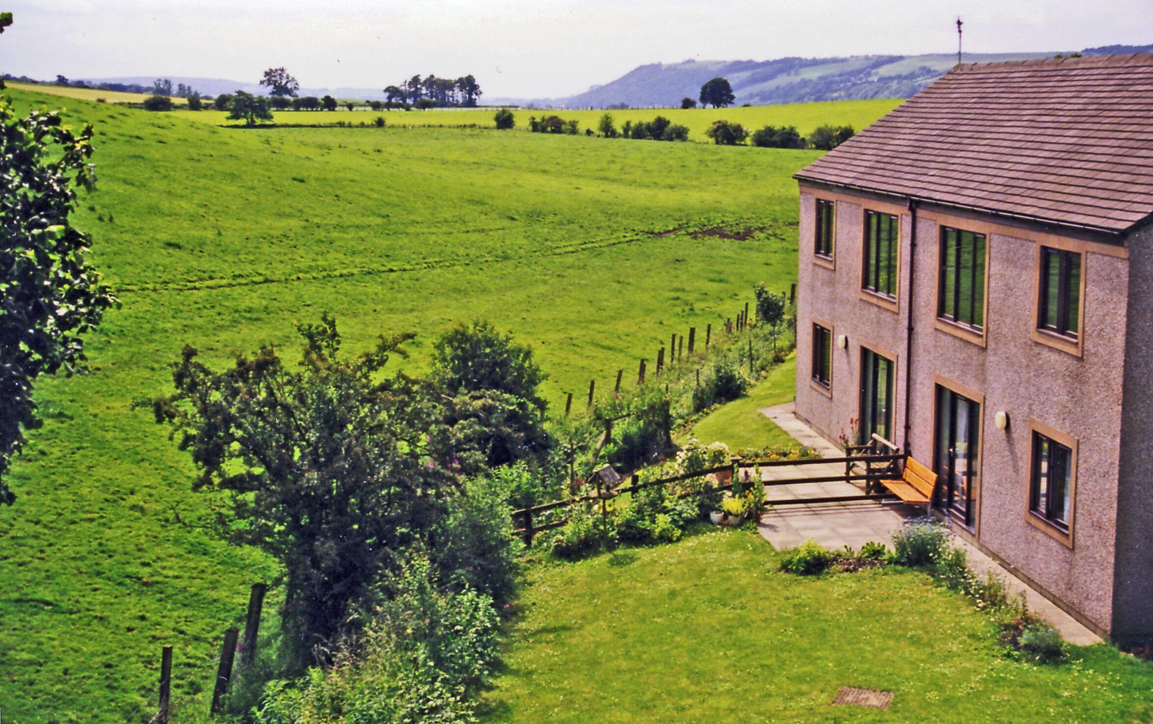 Site of former Hornby station, 1998. View westward, towards Lancaster: ex-Midland (Skipton etc.) - Wennington - Lancaster - Morecambe - Heysham secondary main line. The station was closed 16/9/57 (goods 20/4/64) and even the whole line Wennington - Lancaster (Green Ayre) - Morecambe was closed entirely 3/1/66, leaving a route round via Carnforth as access from Yorkshire to Morecambe and Heysham. Here in 1998 the route is barely recognisable and a modern house has been built where the station was once. The view is south of the village and down the Lune Valley with Crag Lot (1,529 ft.) over on the right.